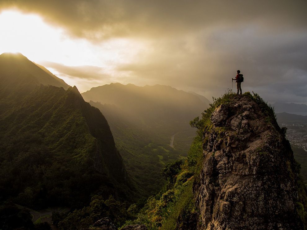 17 Taking a Peak. Photograph by Liz Barney. As beams of light from the setting sun burst through clouds, a triumphant hiker basks in their glow on a summit in Hawaii’s Ko’olau Range.